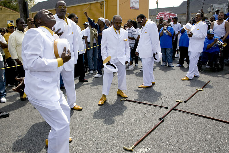 A group of men in white suits and gold shoes perform energetically on a street, holding hats. A crowd watches, and a brass band plays in the background. The scene is lively and festive, with a focus on the performers expressive movements.