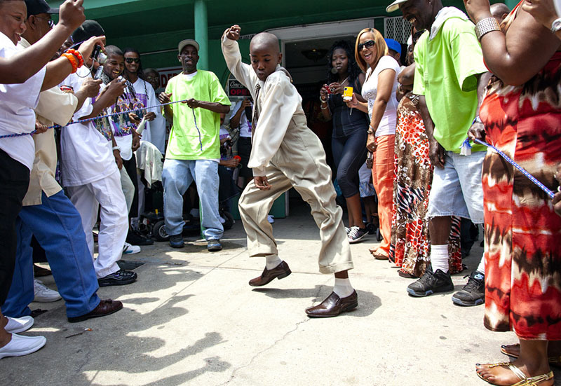 A young boy energetically dances in a beige suit and brown shoes, surrounded by a cheering crowd. The scene is lively, with people casually dressed, many smiling and some taking photos outside a store.