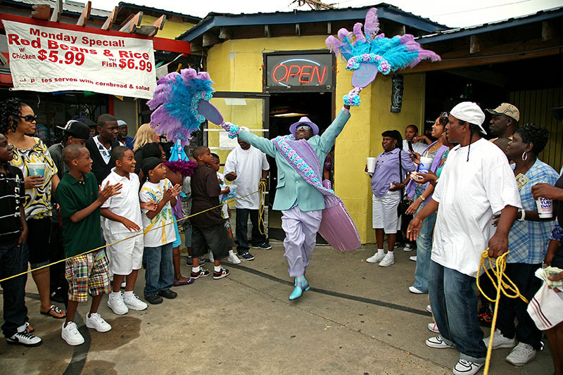 A person dressed in a vibrant costume dances energetically in front of a crowd. The costume features large, colorful feathers and a patterned sash. Onlookers, including children, gather around. A sign advertises food specials in the background.