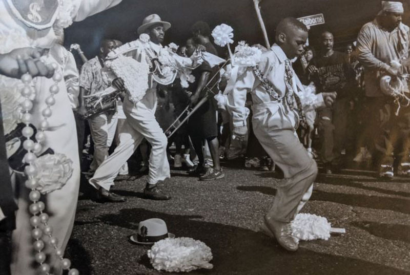 A lively street scene with people dancing in white and decorative attire, holding musical instruments like a trombone. A hat lies on the ground. The energy is festive, with a crowd surrounding the performers, appearing to enjoy the celebration.