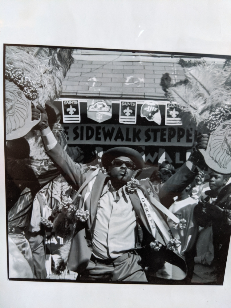 Black and white photo of a person in a suit and hat, dancing energetically with feathered fans. Around them, others join the celebration. A sign in the background reads SIDEWALK STEPPE. The scene is lively and festive.