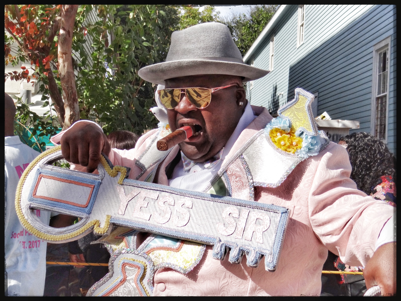 A man in a pink suit, gray fedora, and gold sunglasses bites a cigar while holding a large decorative key with "YESS SIR" embroidered on it.
