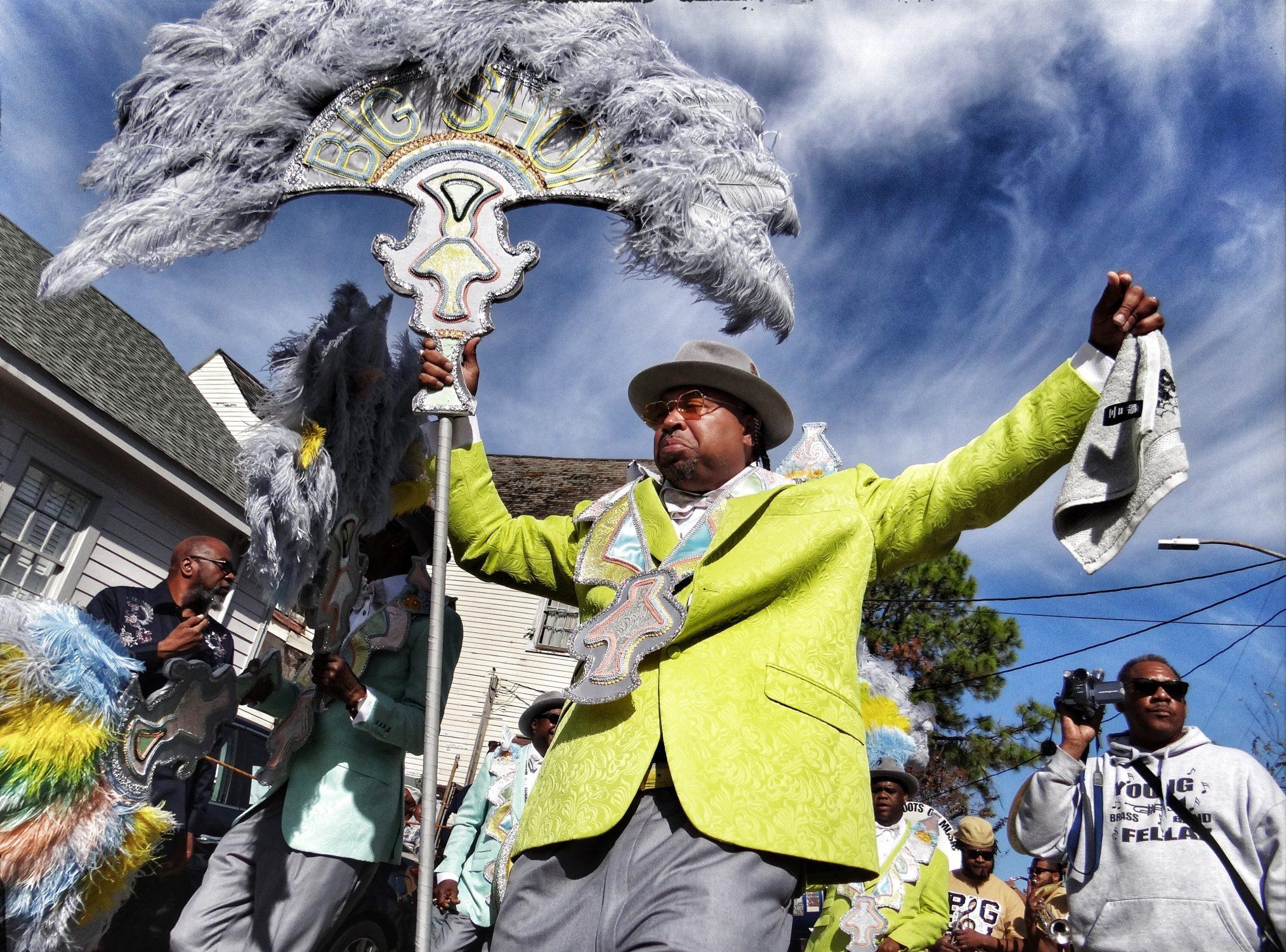 A person in a bright green suit and gray hat leads a vibrant parade, holding a large feathered sign. Surrounding participants wear colorful outfits and carry feathered designs, celebrating under a clear blue sky.