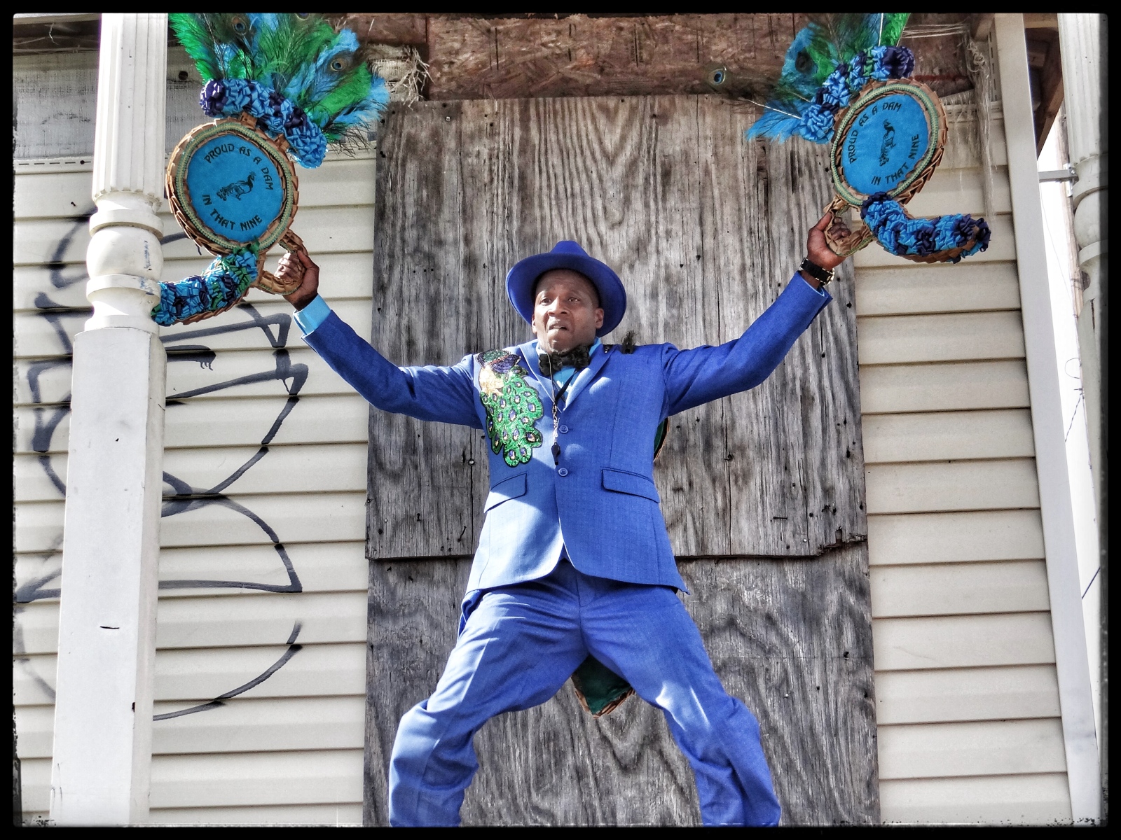 A man dressed in a bold blue suit and hat jumps with his arms wide, gripping two ornate, circular props decorated with peacock feathers and embroidery. Behind him, a worn wooden wall with boarded-up windows and graffiti adds contrast to his vibrant presence.