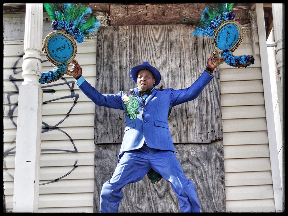 A man in a bright blue suit and hat energetically poses in front of a weathered wooden background. He holds two decorated circular fans with peacock feathers, one in each hand, and has a lively expression. Graffiti is visible on the wall.
