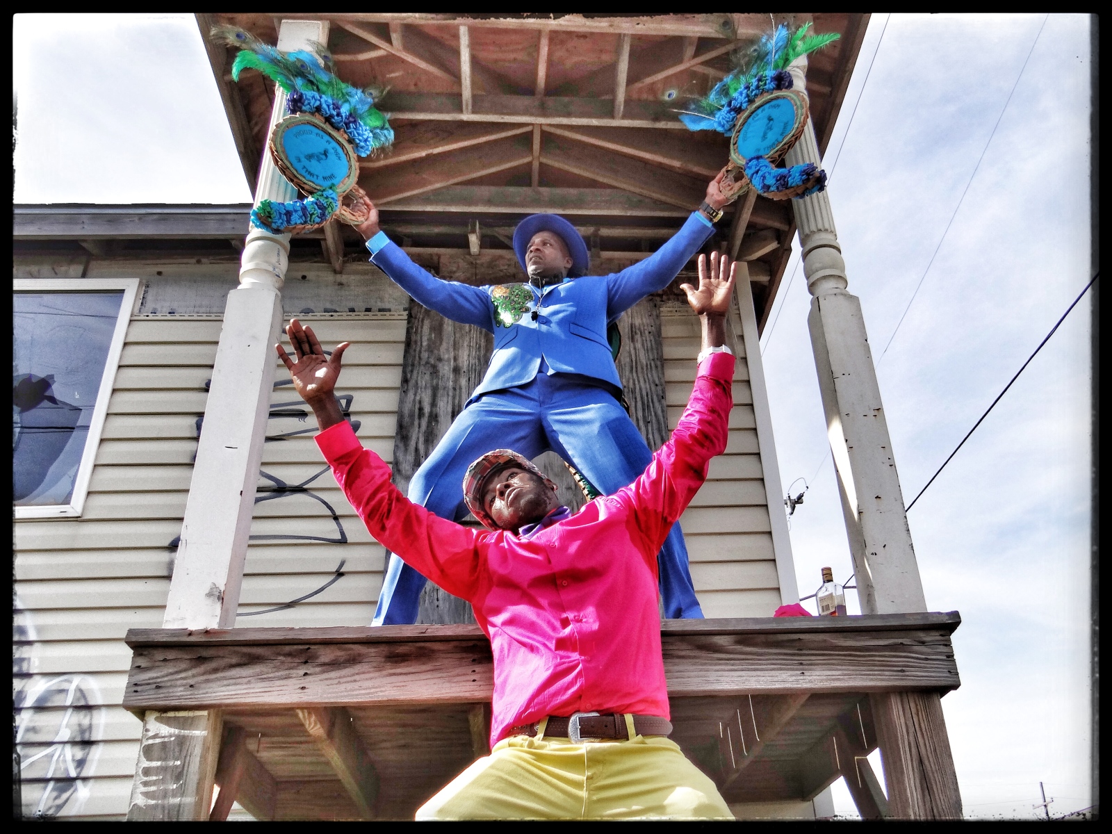 Two men strike dramatic poses on the porch of a weathered house. The man on the upper level wears a blue suit and hat, raising two ornate props adorned with peacock feathers. Below him, a man in a vibrant pink shirt and yellow pants extends his arms upward, mirroring the pose. Graffiti decorates the siding of the boarded up house behind them.