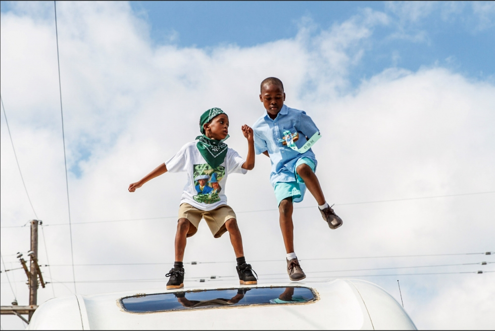 Two children joyfully jump on a white surface against a backdrop of a blue sky with fluffy clouds. One child wears a green bandana and a t-shirt, while the other sports a blue shirt with shorts. Electric poles and wires are visible in the distance.