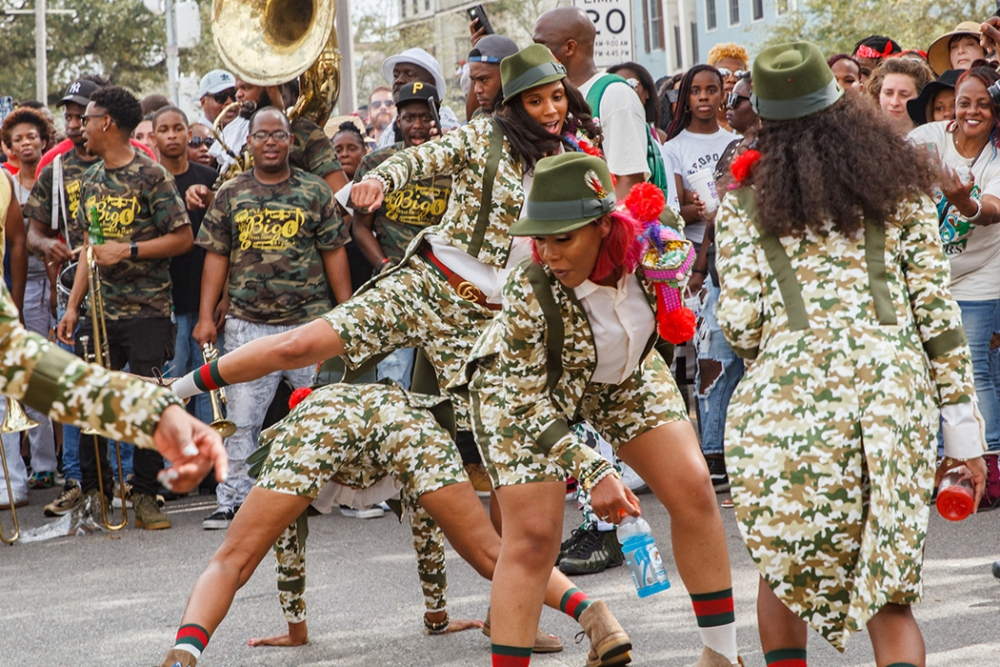 A vibrant parade scene featuring dancers in coordinated green camouflage outfits performing in front of a lively crowd. A marching band member plays a sousaphone, and spectators capture the moment on their phones.