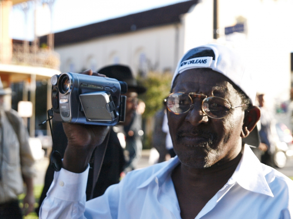 A man wearing glasses, a white shirt, and a cap with New Orleans written on it holds a video camera. He is outdoors in a sunny setting, and there are buildings and other people in the background.