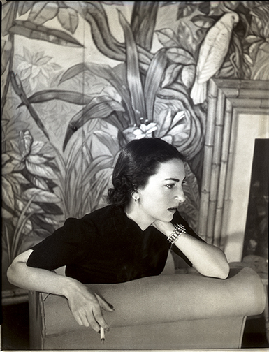 A woman sitting in a chair with a thoughtful expression, holding a cigarette. She wears a dark top and a bracelet. The background features a detailed, tropical mural with large leaves and a parrot.
