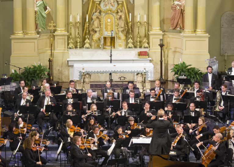 An orchestra performs in a grand hall with a conductor leading musicians playing strings, woodwinds, and brass instruments. The backdrop features an ornate altar with candles and religious decorations.