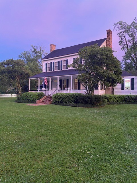 A white, two-story house with a porch and brick chimneys, set in a large grassy yard with surrounding trees. An American flag hangs from the porch, and the sky is a pale purple, suggesting early morning or evening.