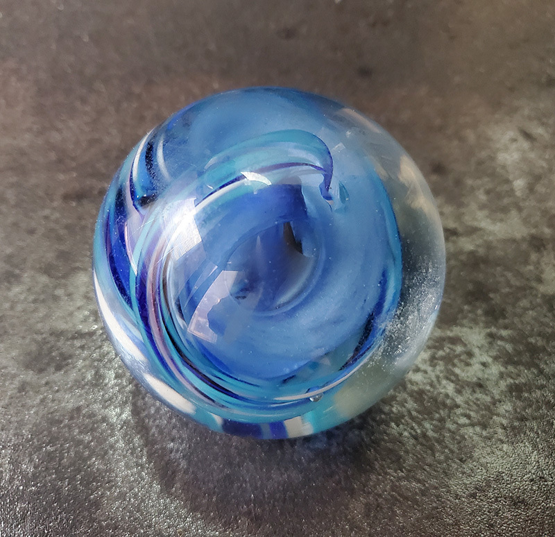 A glass marble featuring swirling shades of blue and hints of turquoise and white, set against a dark, textured background. The marble has a glossy finish and intricate internal patterns.