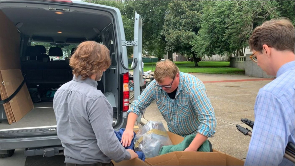 Three people unload items from a van into a cardboard box. One person with sunglasses is sorting items in the box, while the others look on. The van doors are open, and trees are visible in the background.