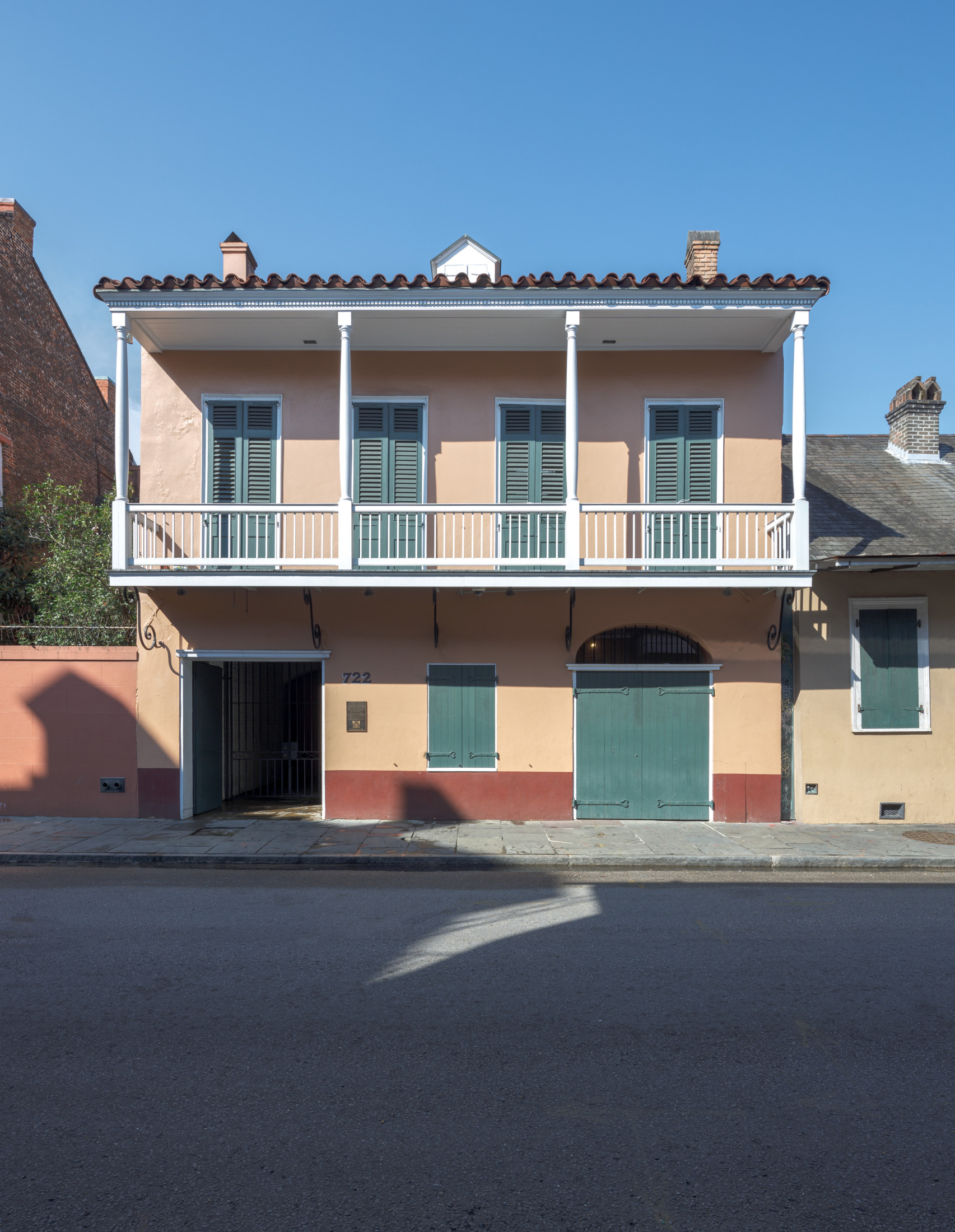 Two-story building with peach-colored facade and green shutters. Balcony with white railings spans the front. Below are arched and rectangular doorways. Clear blue sky above. Quiet street in the foreground.
