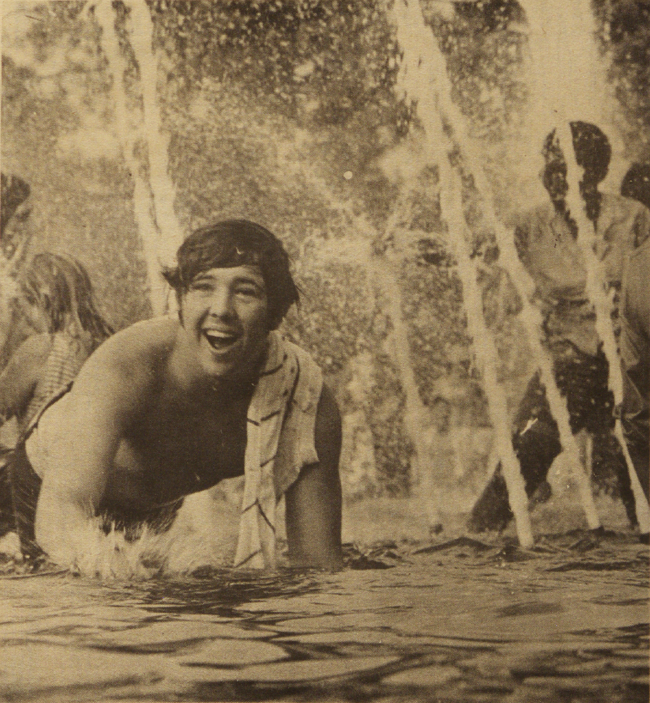 A young person with a joyful expression lies in a fountain, surrounded by water streams, with others in the background. They have a towel draped over their shoulder. The atmosphere is playful and carefree.
