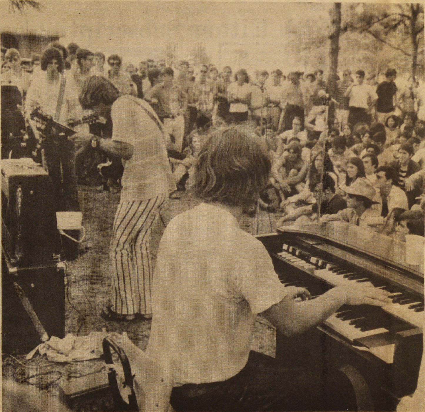 A black-and-white photo of a live outdoor concert. A musician plays the keyboard in the foreground, while another plays guitar. A crowd of people, some seated and others standing, listens intently. Many wear casual 1970s-style clothing.