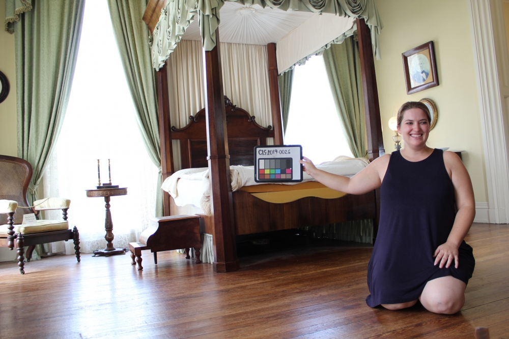 A woman kneels on a wooden floor in a vintage-style room with a canopy bed and ornate curtains. She holds a color calibration chart. The room features classic furniture and framed art.