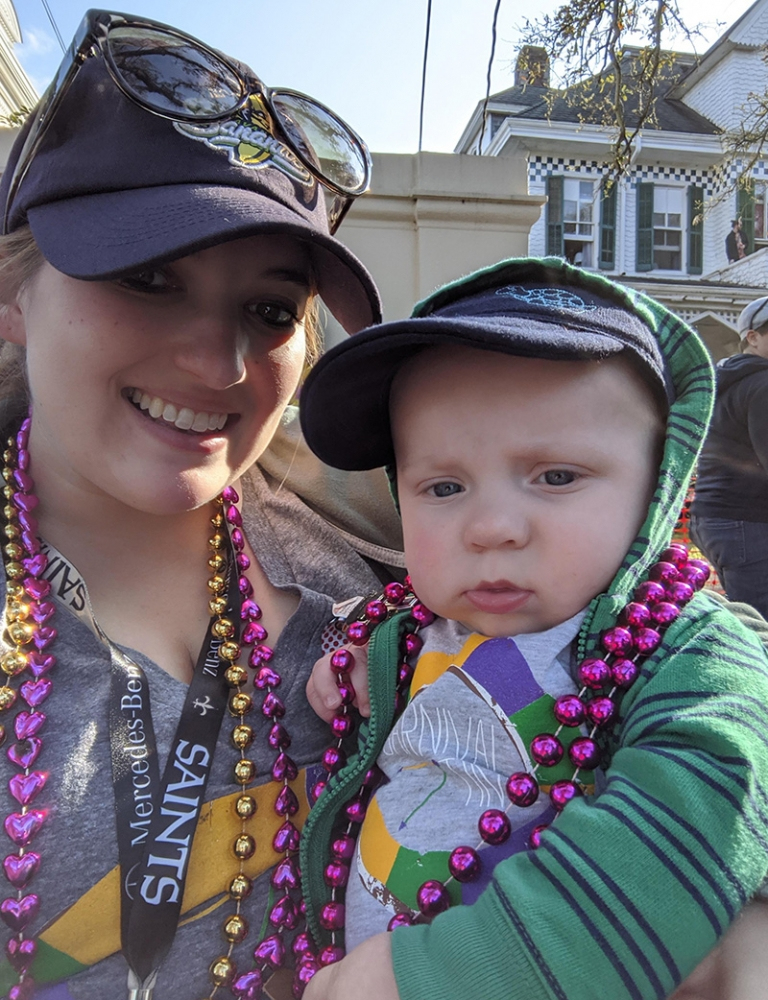 A woman wearing a cap and colorful beads smiles while holding a baby dressed in a hoodie and cap. The baby is also adorned with purple and gold beads. They stand outdoors, and a house is visible in the background.