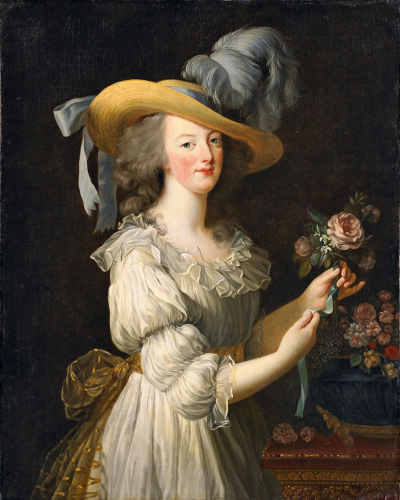 A woman in an 18th-century dress stands with a soft smile, holding a flower. She wears a large hat with a feather and a ribbon. The background is dark, emphasizing her light-colored attire and the floral arrangement beside her.