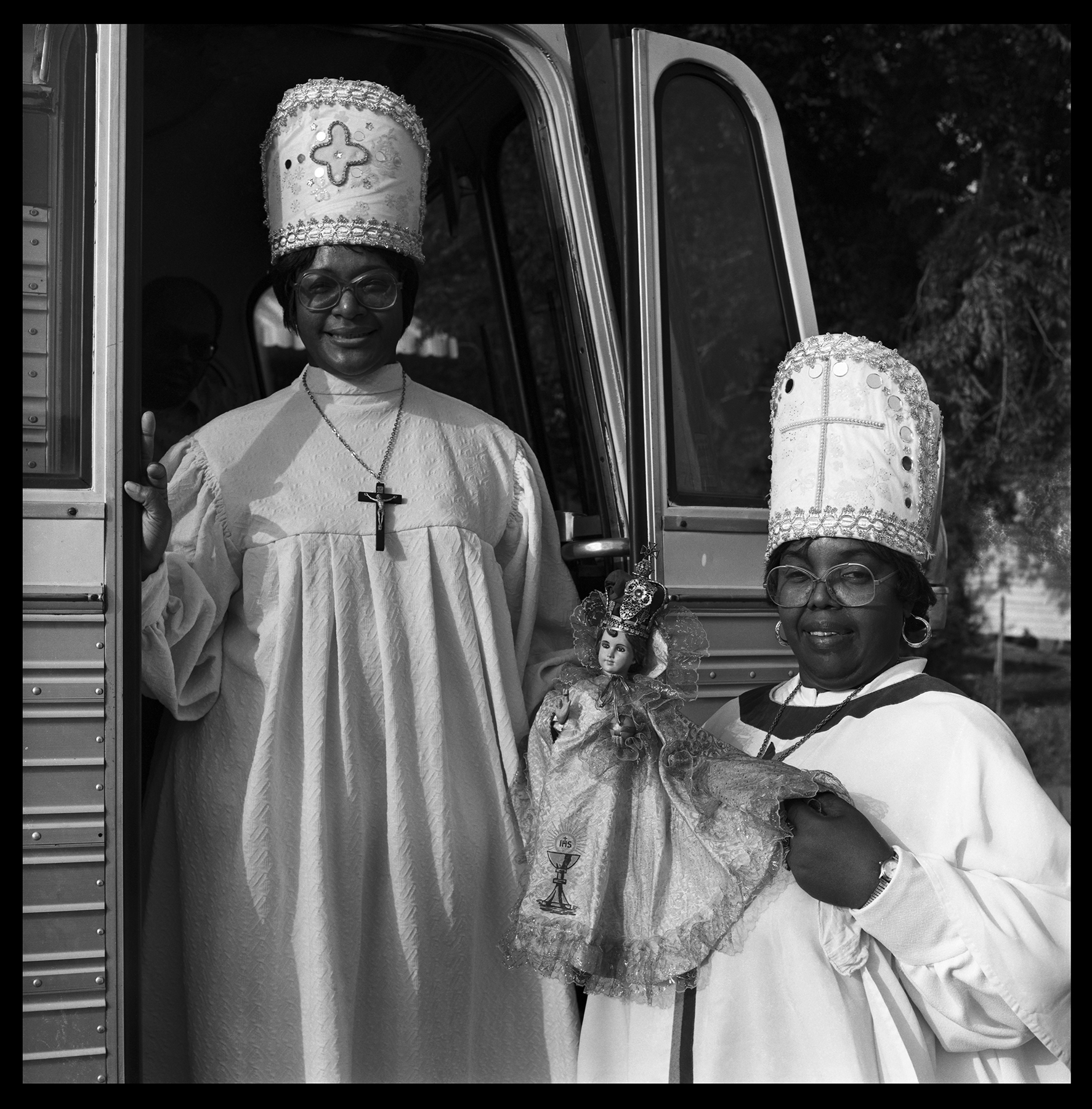 Two people in ceremonial attire stand by a bus. The person on the left wears a cross necklace, and the person on the right holds a doll dressed similarly. Both wear tall, ornate hats. They appear to be participating in a cultural or religious event.