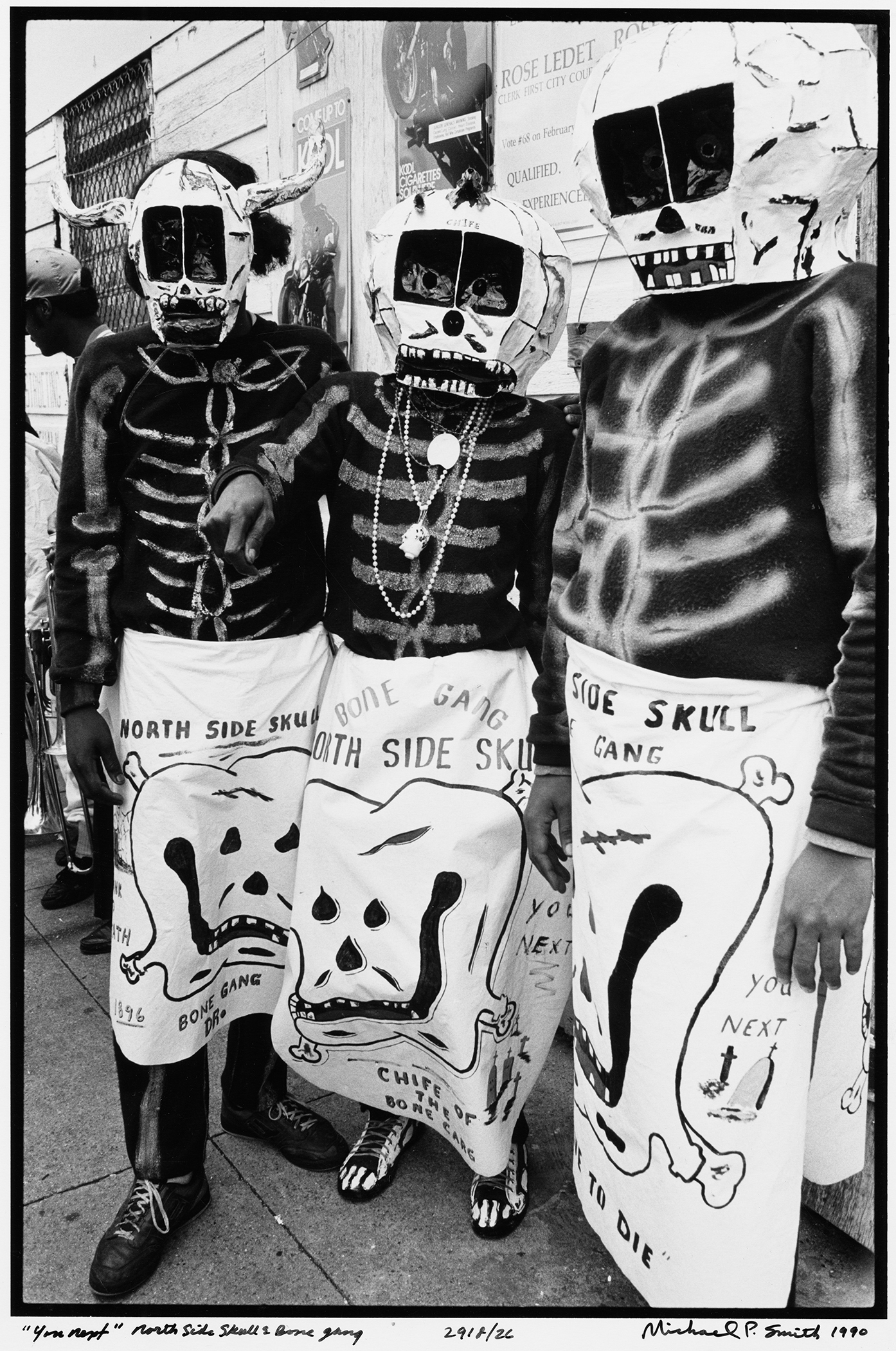 Three people wear skeleton costumes with skull masks and detailed designs on their outfits. They stand side by side on a street with posters on the wall behind them. The image is in black and white.