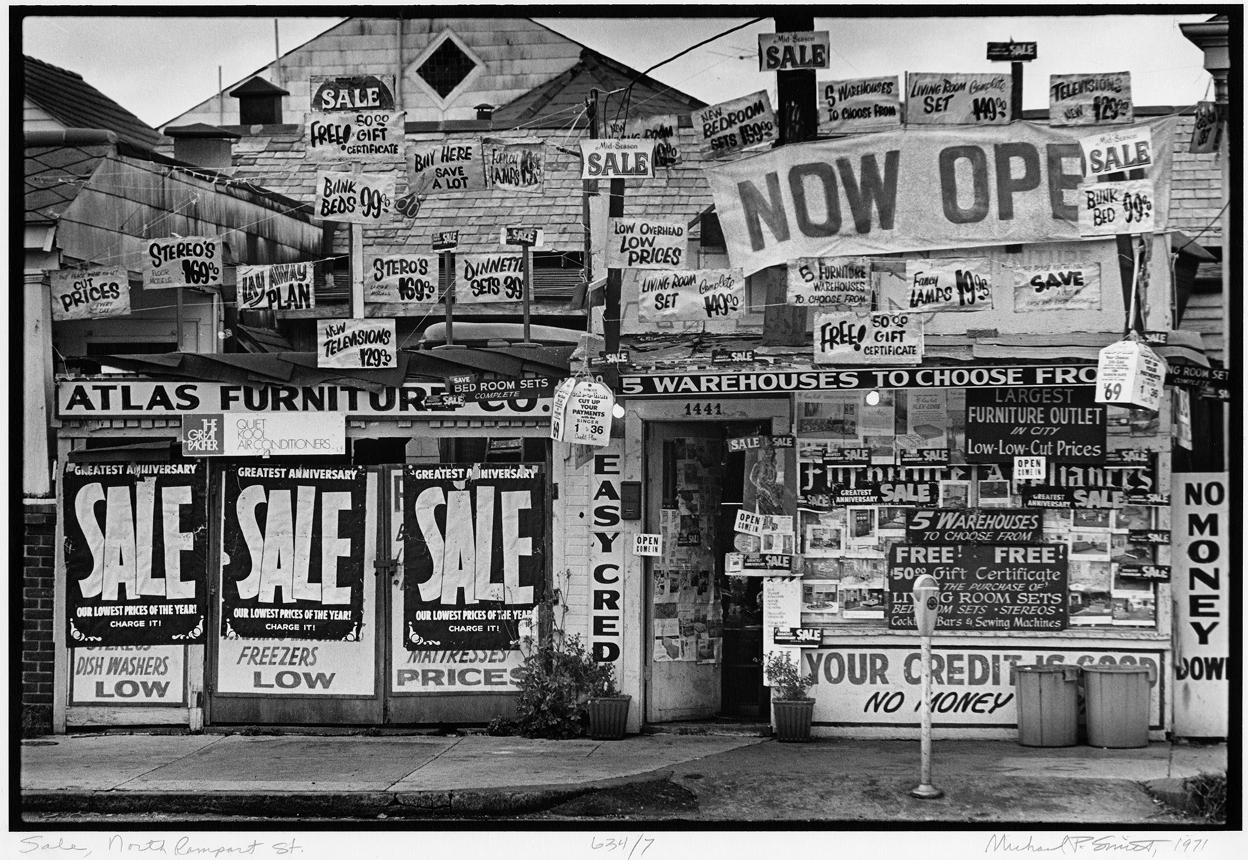 A storefront covered with numerous sale signs advertising discounts on furniture and appliances. Large banners proclaiming NOW OPEN and SALE dominate the display, with various handwritten notices on deals and prices.