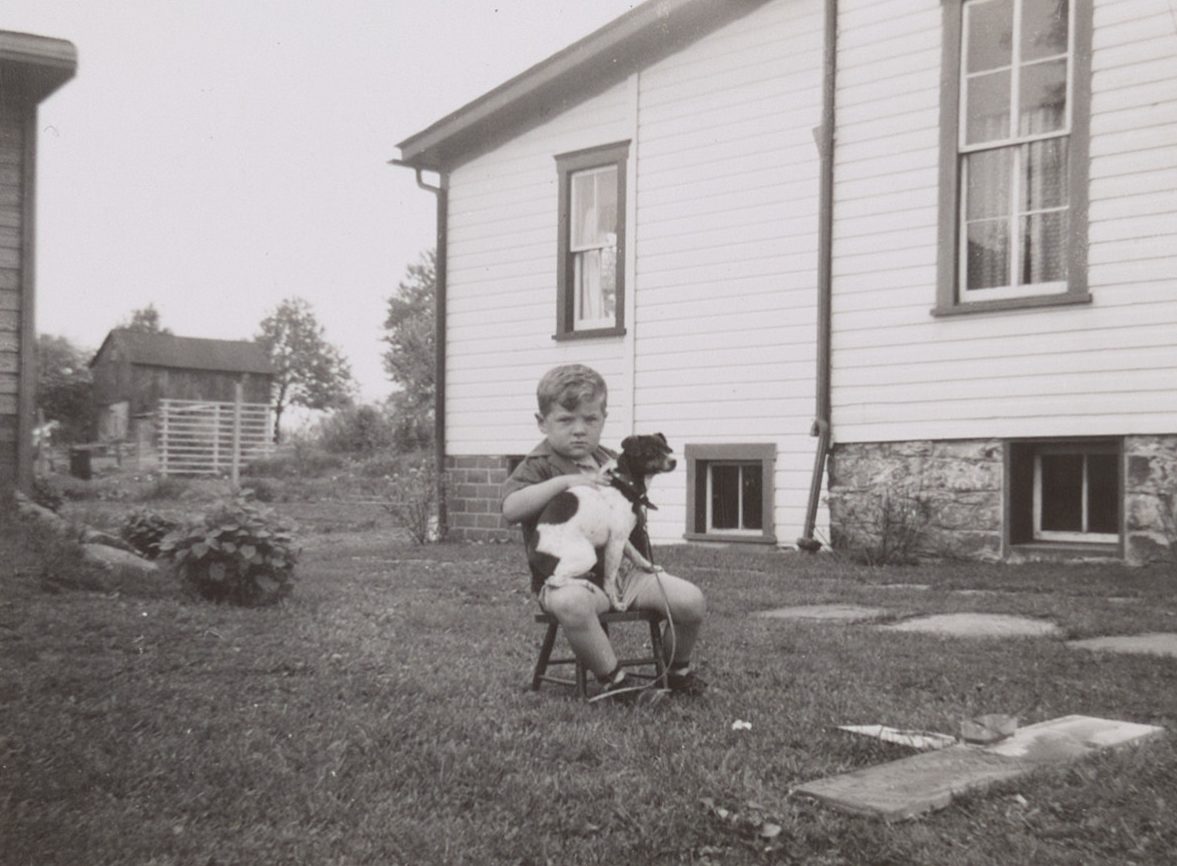 Black and white photo of a little boy siting in a yard with his dog on his lap.