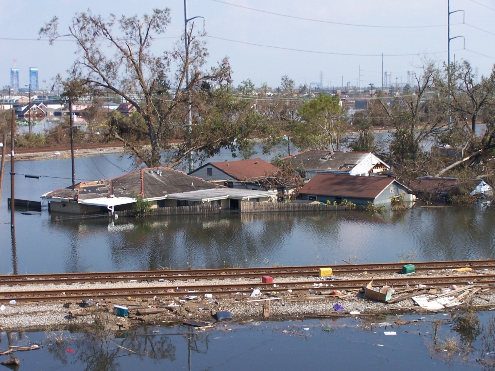 A flooded residential area with submerged houses and scattered debris. Partially submerged trees and power lines are visible. Train tracks run through the foreground, littered with debris. The background shows an industrial skyline.