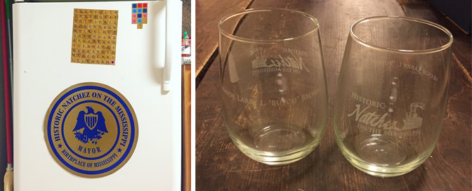 The image shows two parts: on the left is a fridge with a circular Historic Natchez on the Mississippi magnet and a colorful grid magnet. On the right are two clear glasses featuring the same historic logo. They sit on a wooden surface.