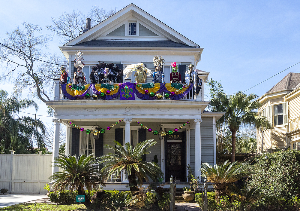 A two-story house is decorated for Mardi Gras with colorful banners, beads, and large masks on the balcony. Palm trees and plants adorn the front yard, and the sky is clear and blue.