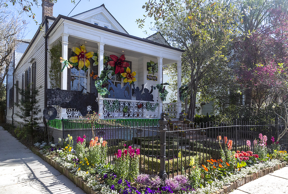 A charming white house with a decorated porch featuring large colorful flowers and silhouettes of musicians. The yard is adorned with vibrant flower beds and greenery, enclosed by a black iron fence, set against a clear blue sky.
