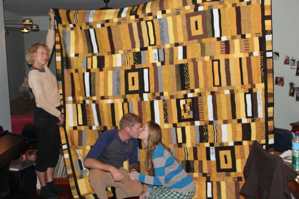 A person holds up a large, colorful quilt with vertical and horizontal patterns in shades of yellow, brown, and black. In front, two people sit closely together, kissing. The room has a cozy atmosphere with dim lighting.