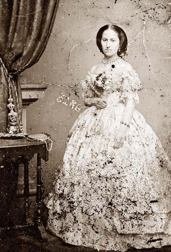 A sepia-toned vintage photograph shows a woman in an elaborate, floral-patterned Victorian dress, standing beside a table with a decorative vase. The image is weathered, with visible scratches and worn areas.