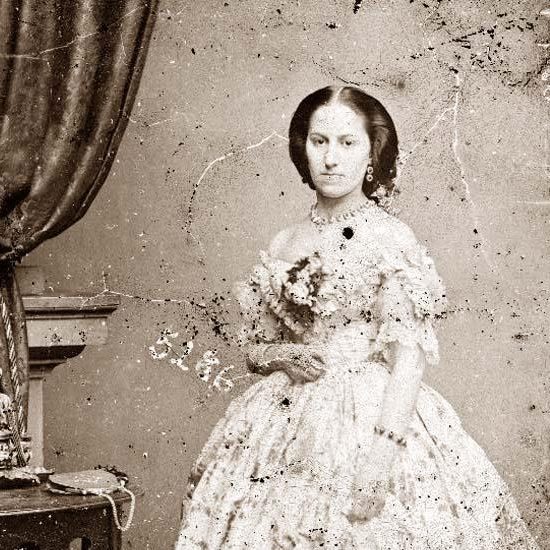 A vintage photograph of a woman in a formal 19th-century dress with lace and floral accents. She is standing next to a small table with a draped curtain in the background. The image is slightly aged and speckled.