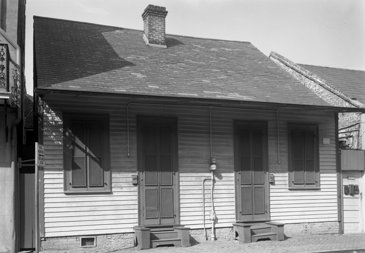 A vintage black-and-white image of a small, wooden house with a steep, shingled roof and a central chimney. Three doors with shutters are visible on the front, and two small steps lead up to the porch. The house sits on a cobblestone street.