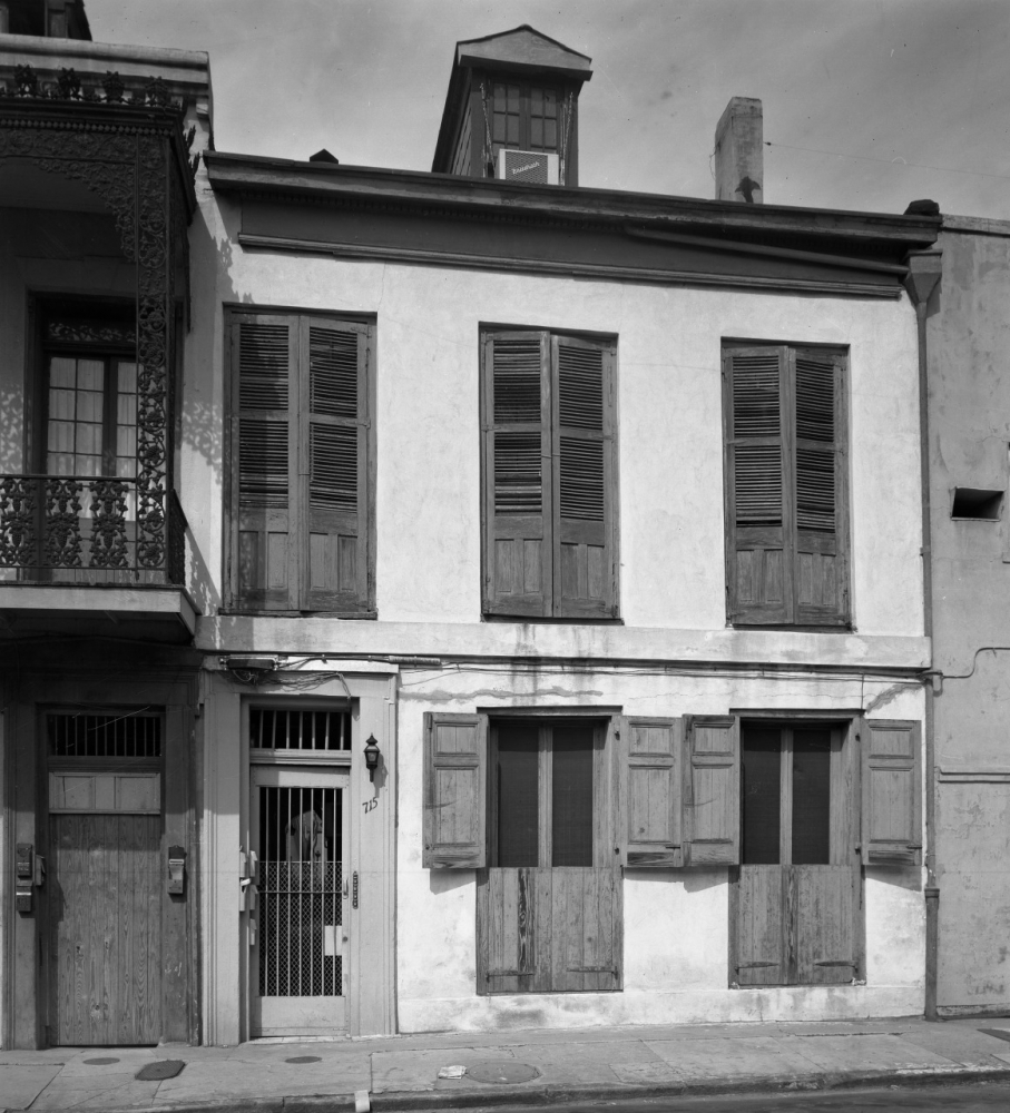A historic two-story building with wooden shutters covers the windows, located next to a decorative wrought iron balcony. The facade is a mix of light and dark tones, with a small porch light next to the entrance door.