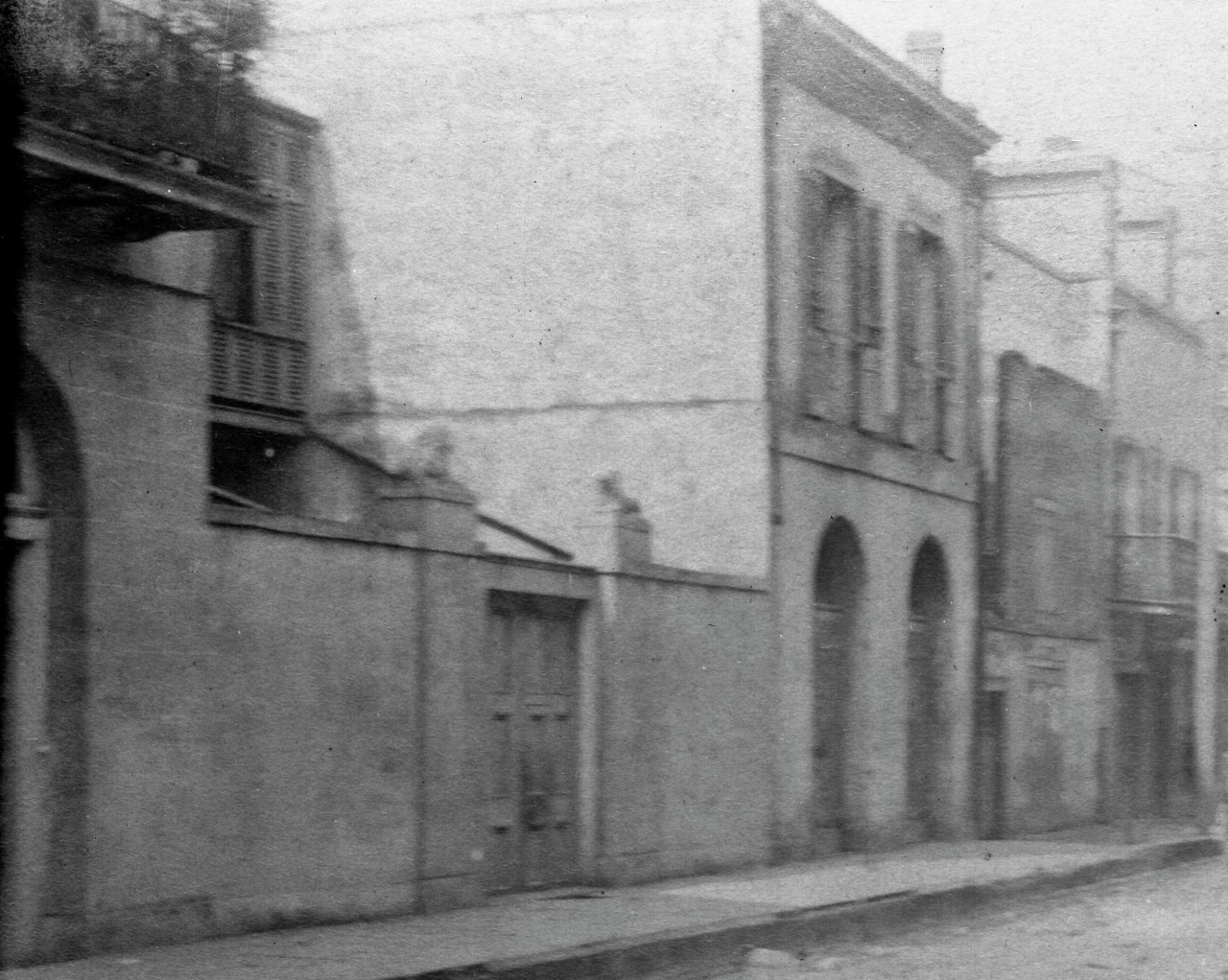 A black and white photo of a narrow, deserted street lined with old buildings. The architecture includes arched doorways and shuttered windows. The scene appears quiet and historical.