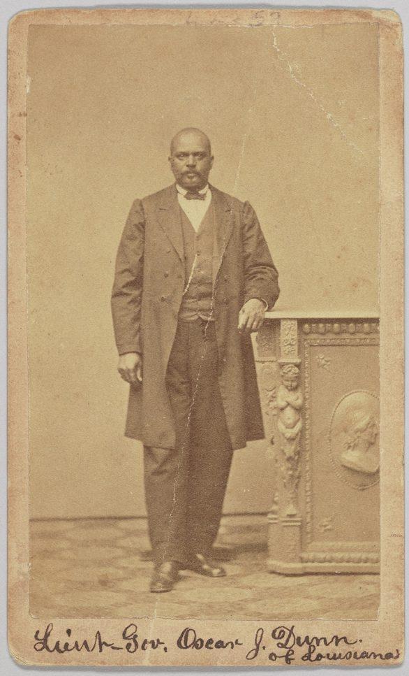 A historical sepia-toned photograph of a man standing beside an ornate table. He is wearing a formal suit and has one hand resting on the table. Handwritten text at the bottom reads Lieut. Gov. Oscar J. Dunn of Louisiana.