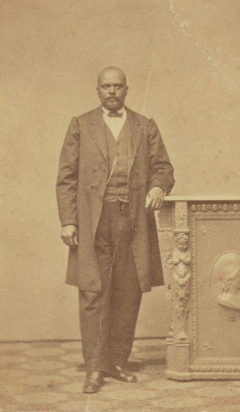 A sepia-toned historical photograph of a bald man with a mustache, wearing a long coat and suit. He stands with one hand resting on an ornate, carved wooden pedestal. The floor has a checkered pattern.