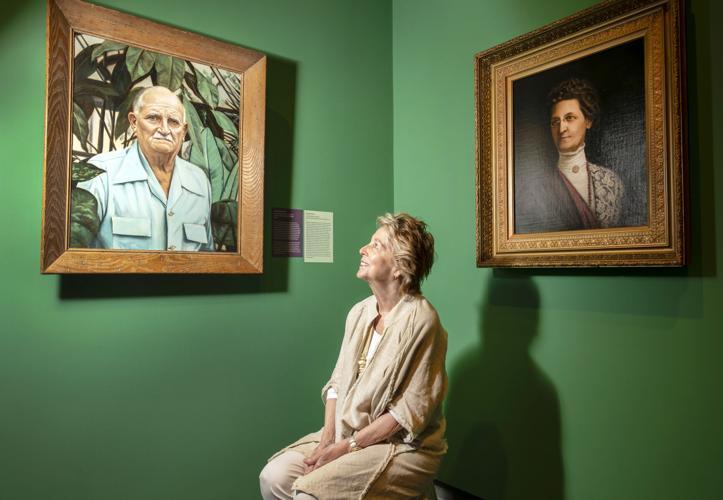 A woman sits on a stool in an art gallery, looking up at two large portrait paintings on a green wall. One portrait depicts a man in a blue shirt, and the other features a woman in historical attire. The gallery lighting highlights the artworks.