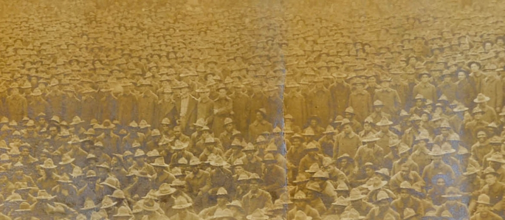 A sepia-toned historical photograph showing a large crowd of people, mostly men, wearing hats and coats, gathered closely together. The image has a slightly faded and rustic appearance.