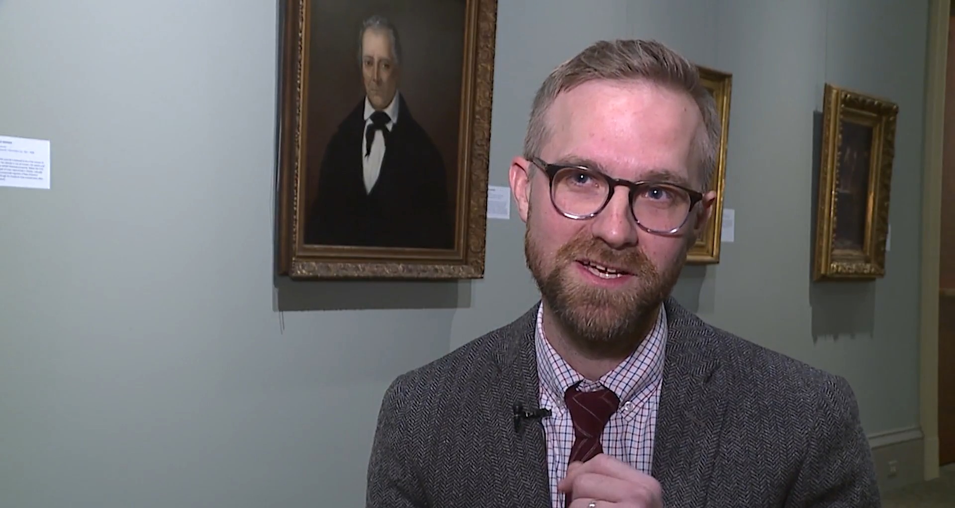 A man being interviewed, standing in front of art portraits at a museum. He is wearing a grey suit jacket, dark tie, and patterned dress shirt.