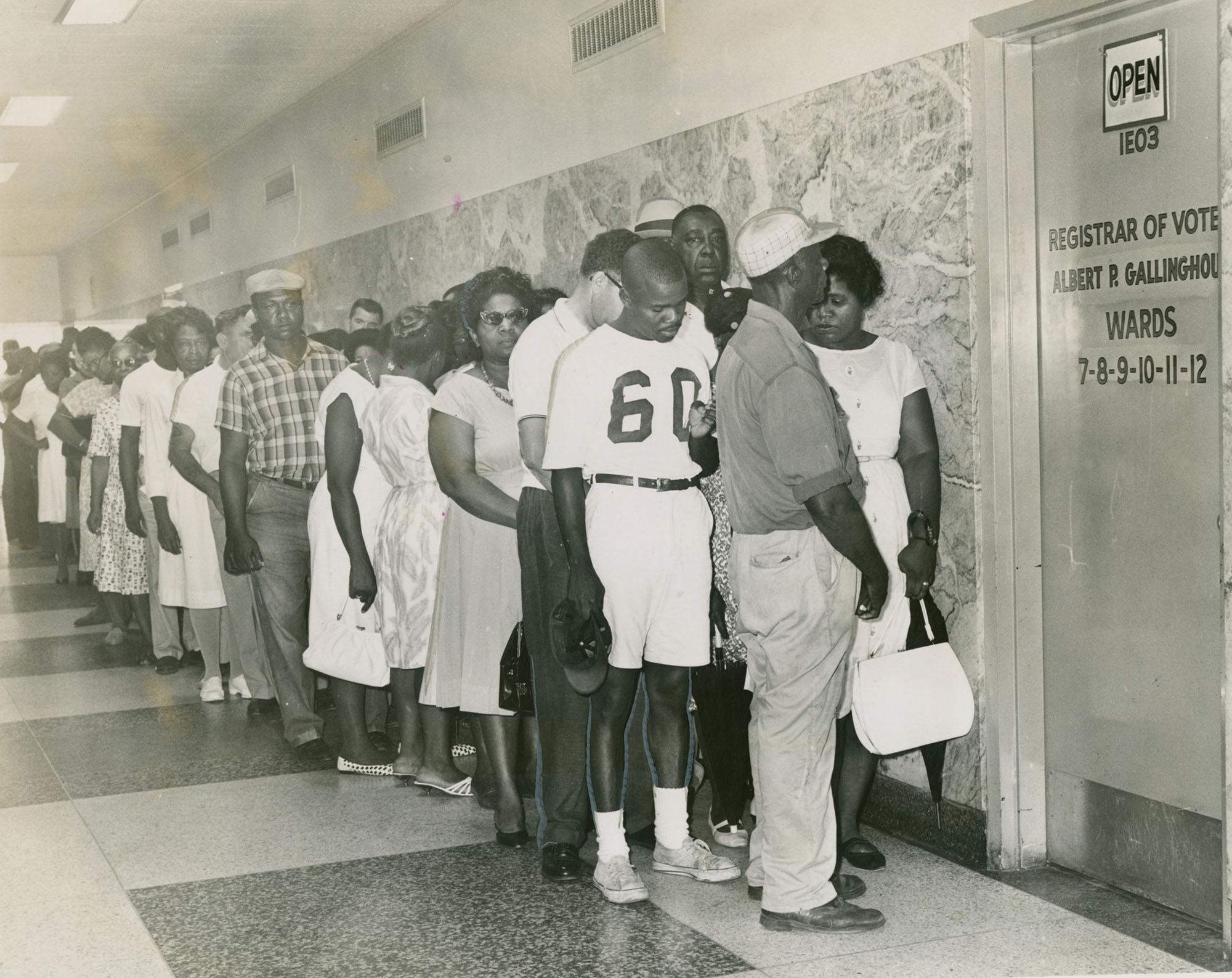 A long line of people, mostly African American, stand in a hallway waiting to access the Registrar of Voters office. A sign on the door indicates its open for wards 7-12. The image captures a moment of civic engagement.