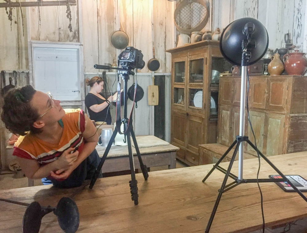 Two people set up filming equipment in a rustic kitchen. One adjusts a camera on a tripod while the other works near a vintage cabinet. A ring light is positioned on a wooden table in the foreground.