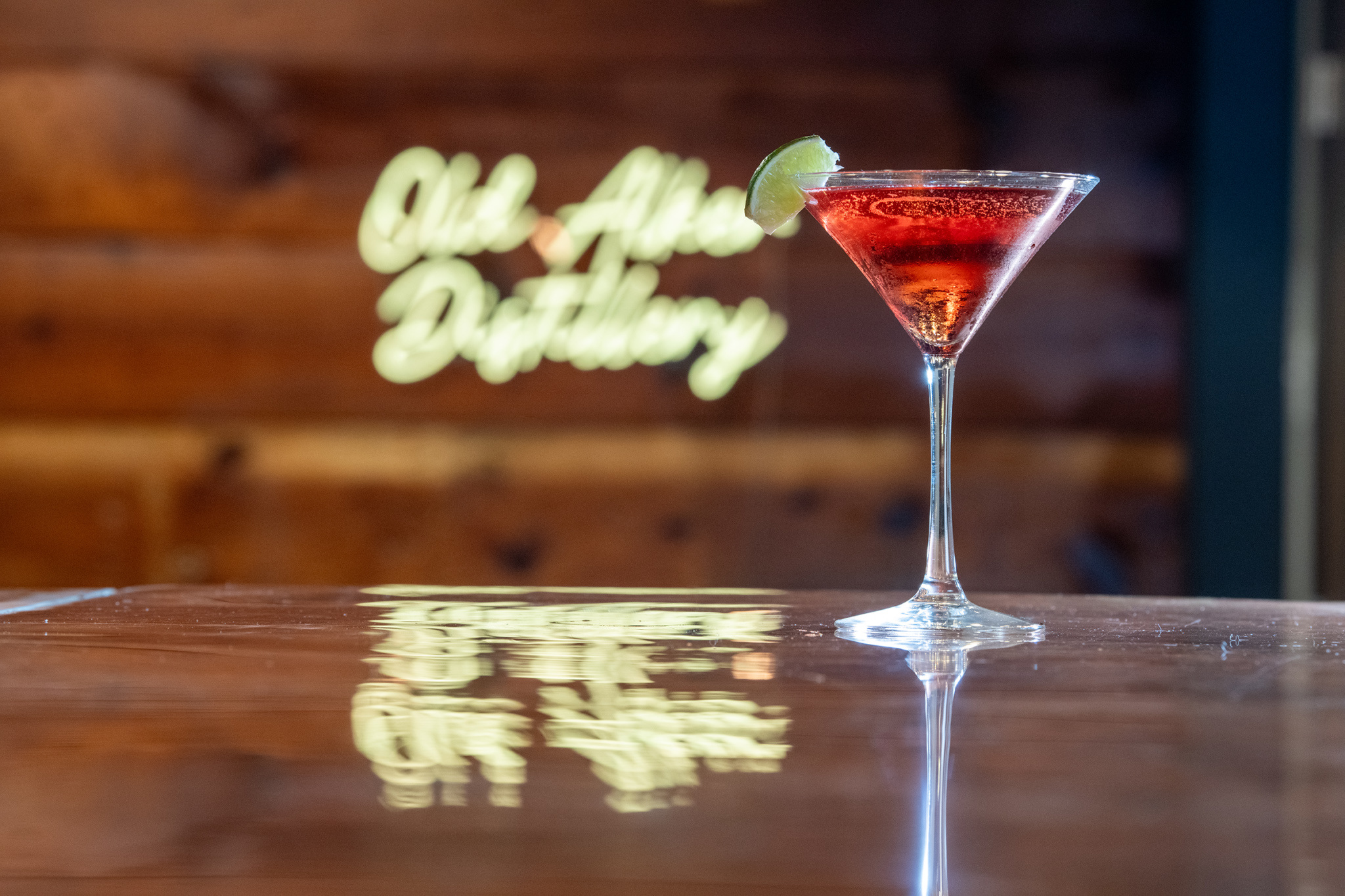 A cocktail in a martini glass with a lime wedge sits on a shiny bar surface. The background features a blurred sign reading Old Adobe Distillery against a wooden wall.