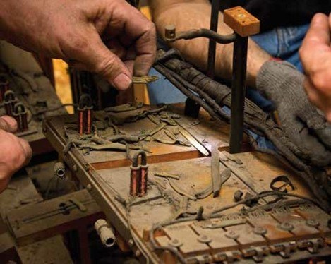 Close-up of hands repairing or assembling a complex mechanical device with gears, levers, and small metal components. People are wearing gloves and using tools. The device appears intricate and machine-like.