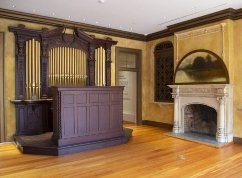 A vintage organ with tall pipes stands in a room with wooden flooring and brown walls. Theres a decorative fireplace with an ornate mantel and a painting above it on the right wall.
