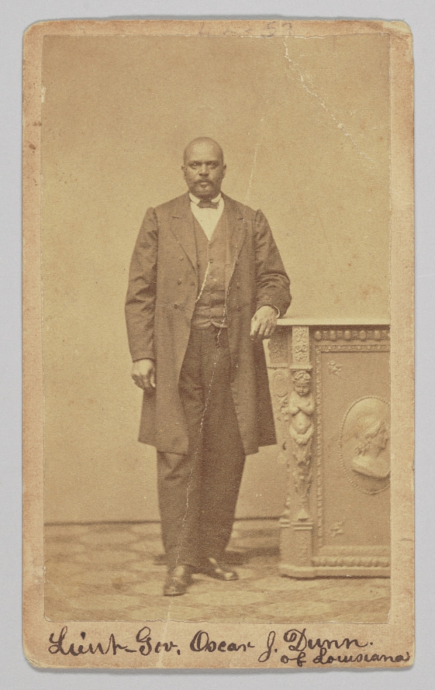 A historical sepia-toned photograph of a man standing next to a carved pedestal. He is dressed in formal attire with a long coat and bowtie. Handwritten text below reads Lieut. Gov. Oscar J. Dunn of Louisiana.