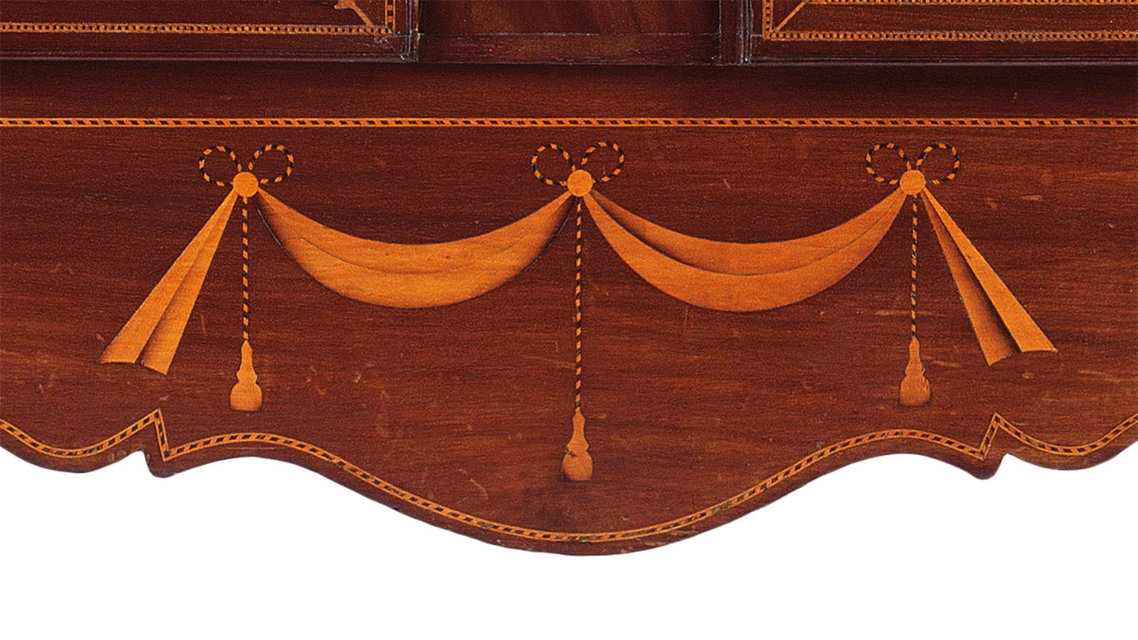 Intricate wood inlay design on a dark wooden surface featuring symmetrical swags and tassels crafted in lighter wood with dotted accents. The edges are scalloped with delicate ornamental details.